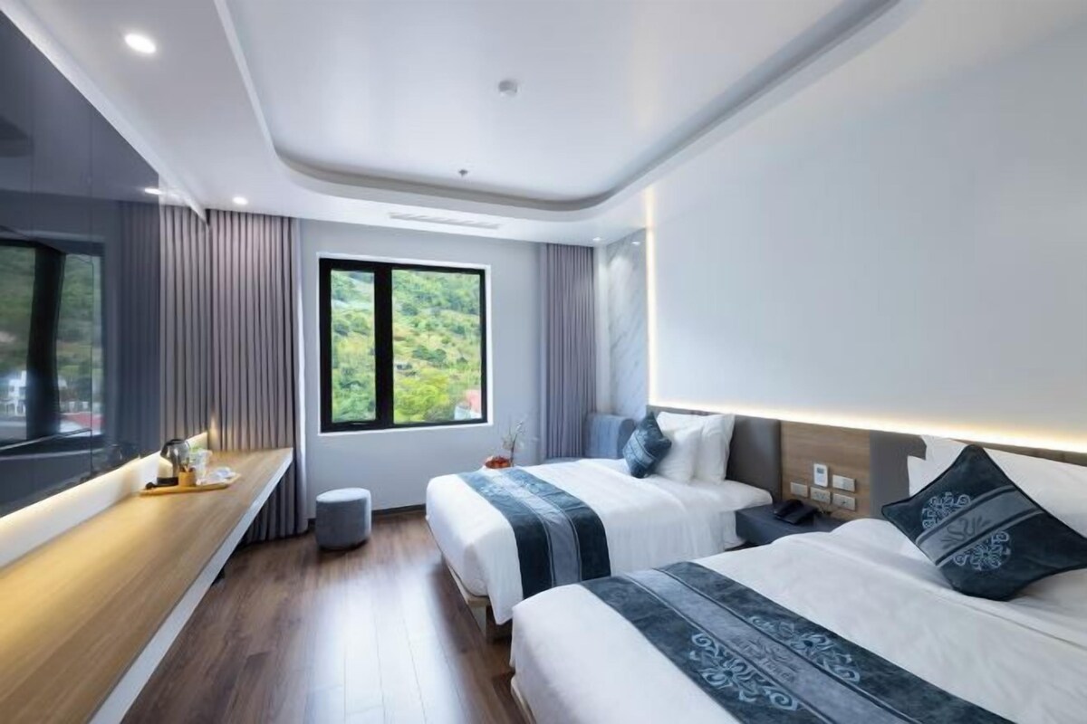 A modern hotel room with two beds, a wooden bedside table, a large window with a view of greenery, and a built-in bench along the window.