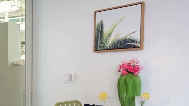 A small dining area with a white table, two green chairs, a vase with pink flowers, and a framed picture on the wall. - Ocean View Studio • by Magen's • Pool & Generator (Northside)