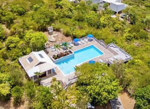 An aerial view of a property with a swimming pool surrounded by lounge chairs and umbrellas, a covered patio area, and a building with a sloped roof.
