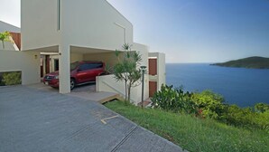 A modern building with a red car parked in the driveway, a clear view of the sea, and a hilly landscape in the background. - Ocean View Studio • by Magen's • Pool & Generator (Northside)