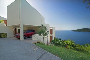 A modern building with a red car parked in the driveway, a clear view of the sea, and a hilly landscape in the background. - Ocean View Studio • by Magen's • Pool & Generator (Northside)