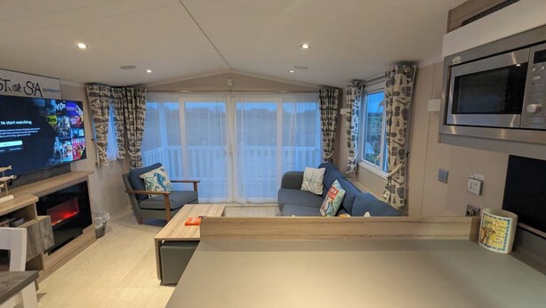 A modern living room with a flat-screen TV, a fireplace, a sofa set, and a large window with curtains. - Lighthouse Lodge Dog Friendly Nr Sutton on Sea (Sutton on sea)