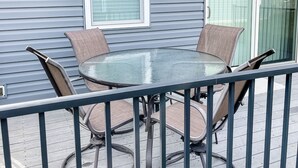 Une terrasse avec une table en verre et quatre chaises, une rambarde et une fenêtre avec des stores.