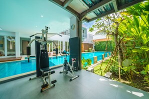 Un gimnasio con equipo de ejercicio y una piscina, rodeado de vegetación.