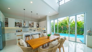 A modern kitchen with white cabinets, a wooden dining table, wicker chairs, and a view of a swimming pool. - Villa Ammanya (Canggu)