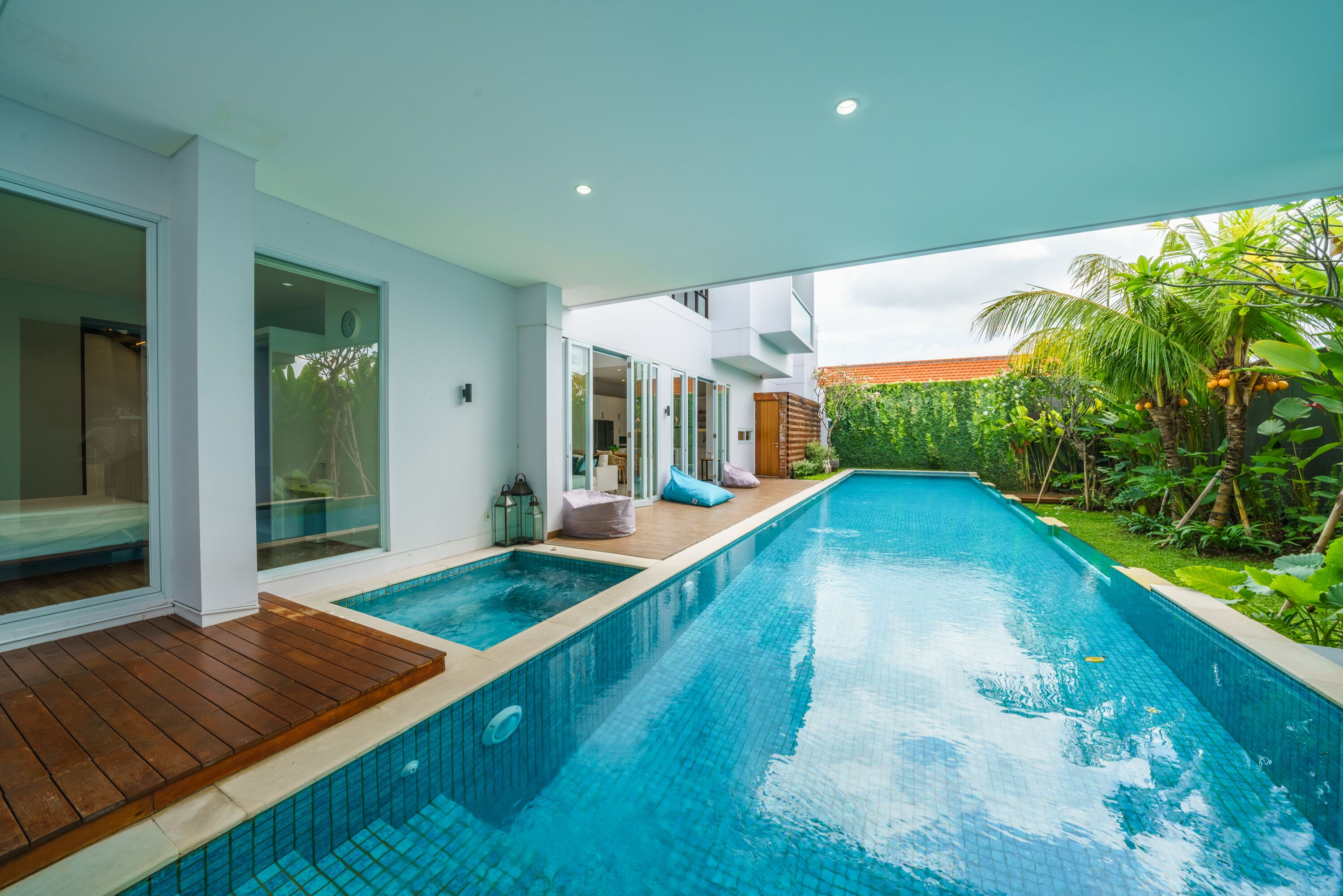 A modern pool area with a wooden deck, glass doors, and a view of lush greenery.