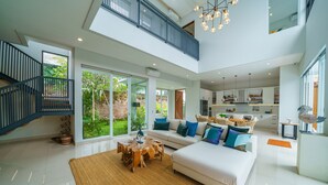 A spacious living area with a high ceiling, a staircase, and a kitchen in the background. - Villa Ammanya (Canggu)