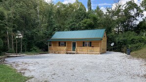 A wooden cabin with a green roof, a gravel driveway, and a forested backdrop. - Peaceful cabin in the center of riding country!  (Duff)