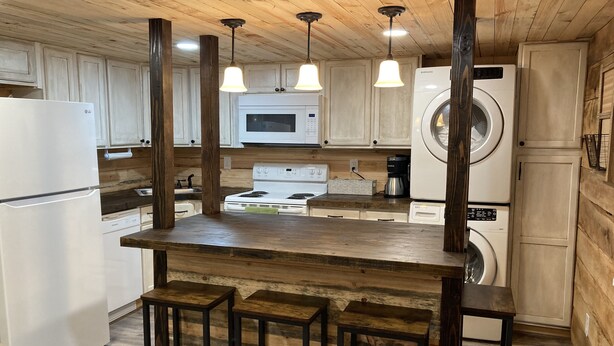 A kitchen with wooden cabinets, a central island with stools, a microwave, a stove, and a washing machine. - Peaceful cabin in the center of riding country!  (Duff)