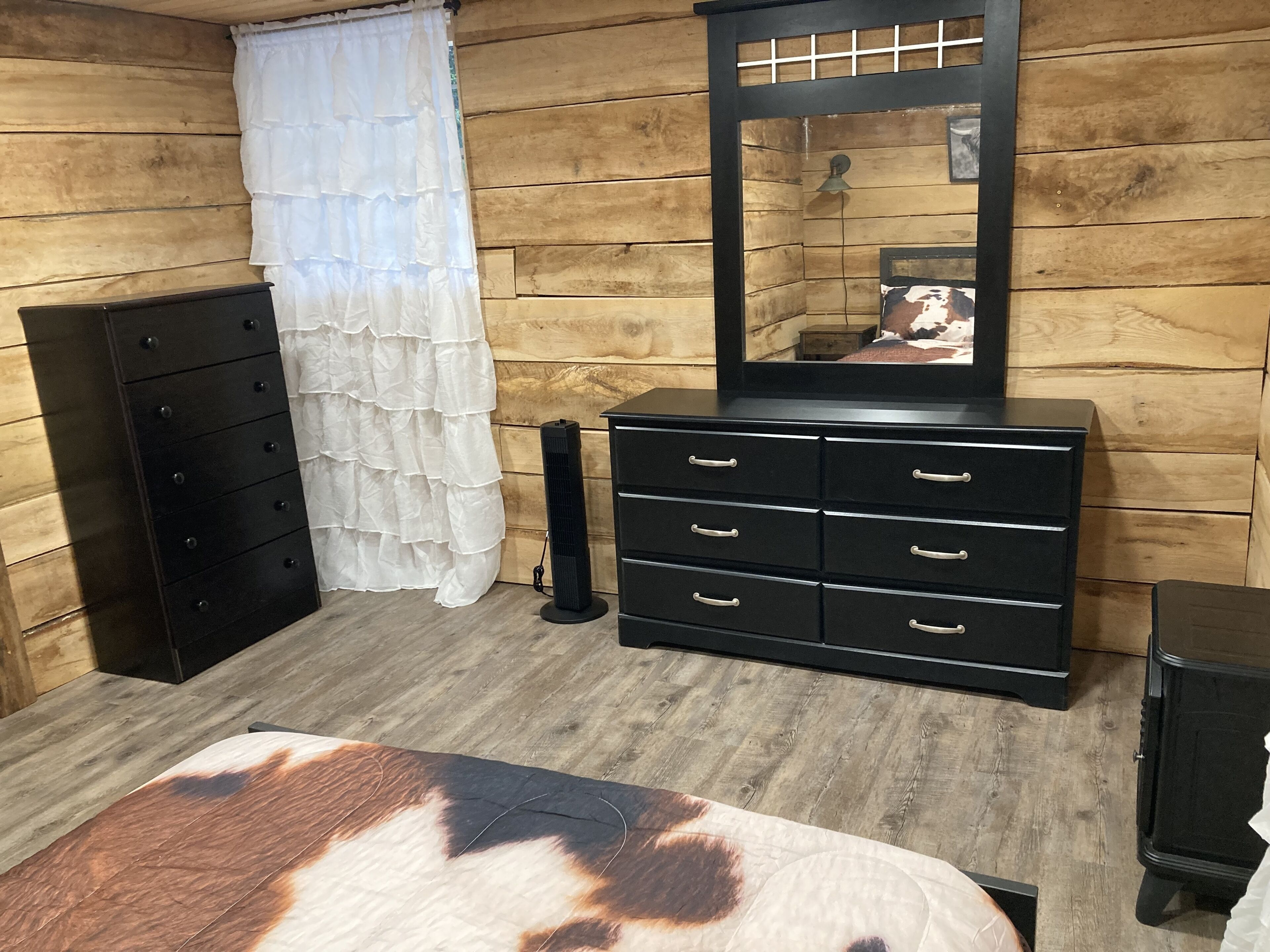 A room with wooden walls, a black dresser with multiple drawers, a mirror, a bed with a cow print blanket, and a white curtain.