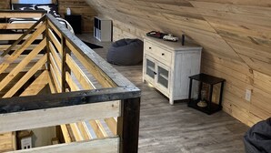 A loft bedroom with a sloped ceiling, wooden beams, a bed with a cow print bedspread, a dresser, a small table, and a bean bag chair. - Peaceful cabin in the center of riding country!  (Duff)