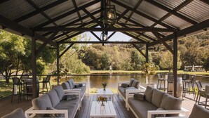 A covered outdoor seating area with sofas and tables, overlooking a lake and surrounded by trees. - Ampersand Estates (Peerabeelup)