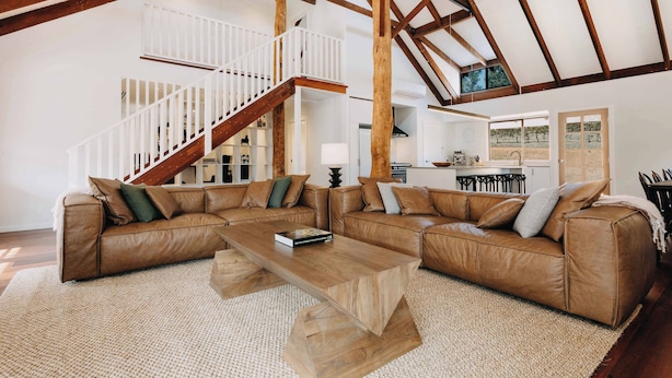A spacious living area with a large L-shaped brown leather sofa, a wooden coffee table, and a white staircase with a railing. - Ampersand Estates (Peerabeelup)