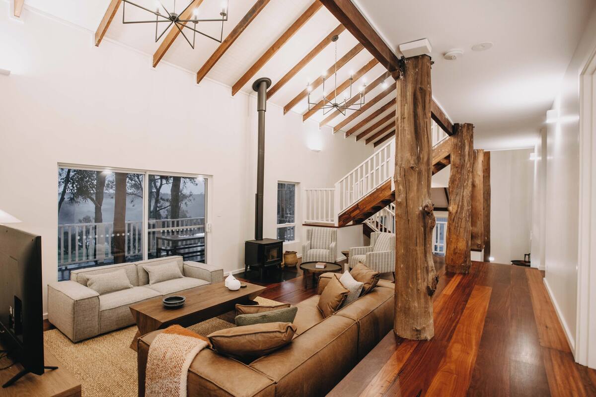 Amplia sala de estar con un sofá grande, una mesa de centro y una chimenea. Se observa una escalera con vigas de madera y vistas al exterior a través de amplios ventanales.