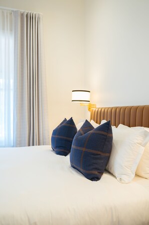 A hotel room with a bed, pillows, a headboard, a lamp, and a window with curtains. - Ampersand Estates (Peerabeelup)