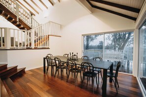 A dining area with a wooden table and chairs, a staircase with white railings, and a large window with a view of trees and a white fence. - Ampersand Estates (Peerabeelup)