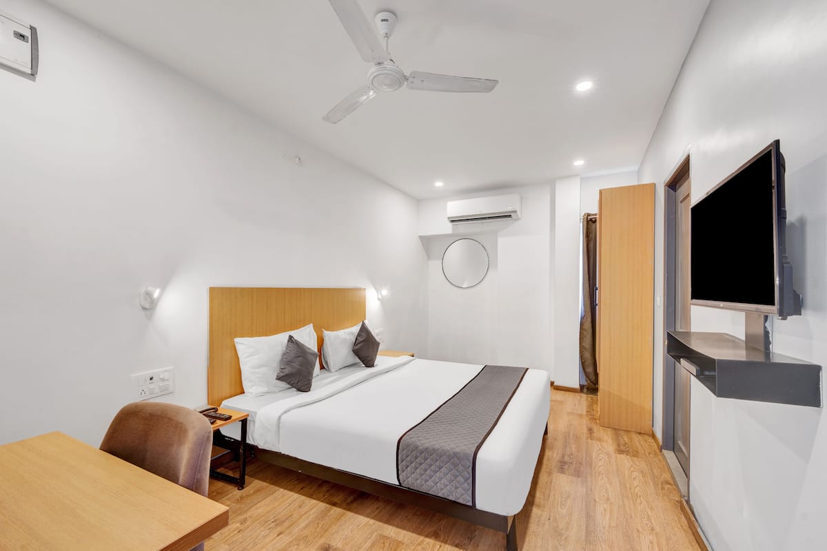 A modern hotel room with a bed, a desk, a chair, a ceiling fan, an air conditioning unit, a television, and a wooden door.