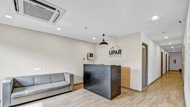 A hotel reception area with a grey sofa, a reception desk, and a logo on the wall. - UPAR Hotels Uthandi ECR (Chennai)