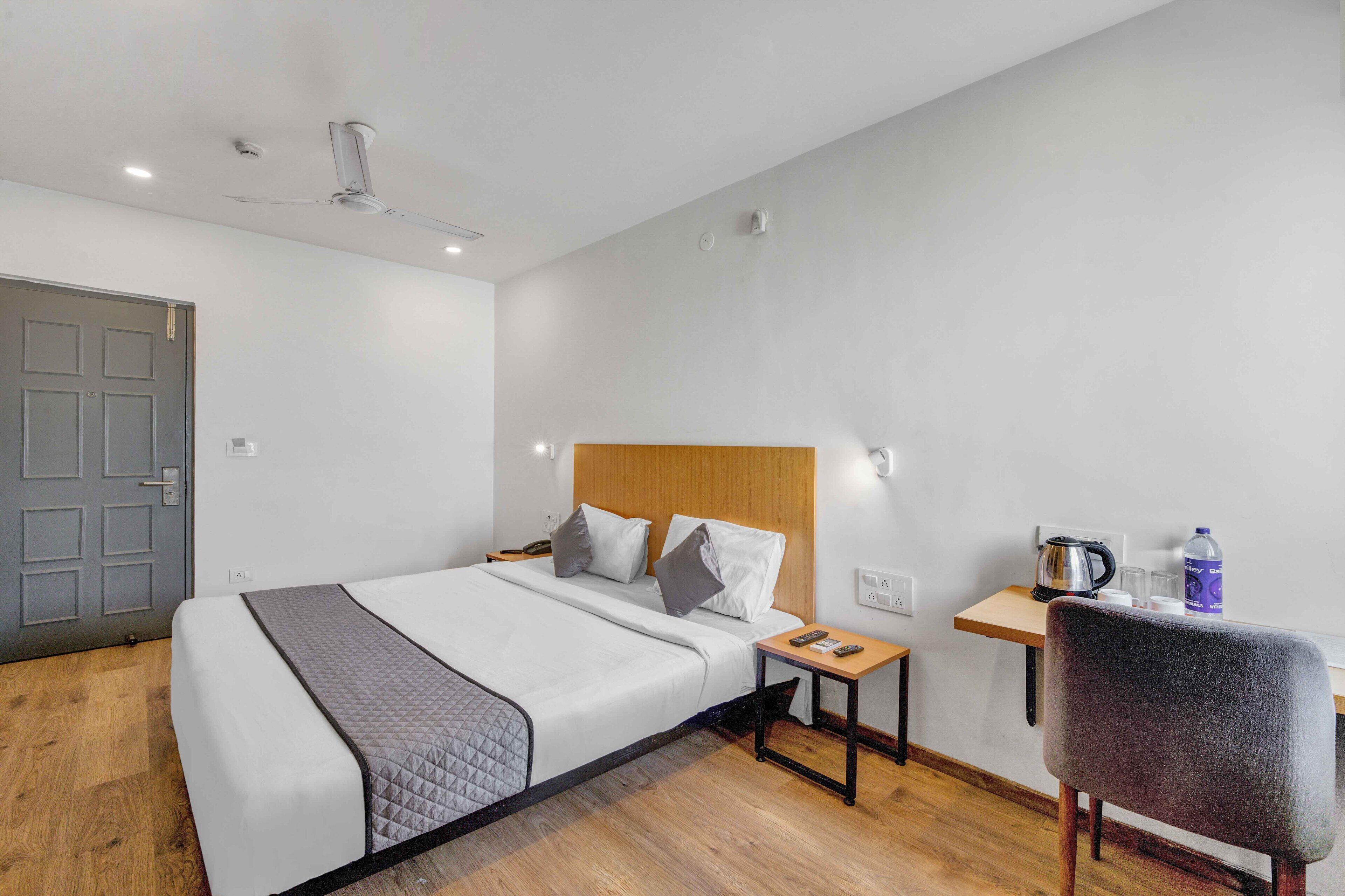 Habitación de hotel con cama, mesita de noche, silla y un escritorio pequeño con tetera y tazas.
