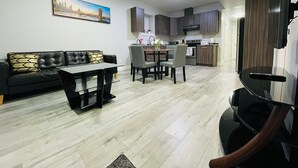 A modern living room with a black leather sofa, a glass coffee table, and a dining area with a small table and chairs. The kitchen features dark wooden cabinets and a stainless steel range hood. - Stylish, clean&cozy new 2 bedroom guest suite (Abbotsford)