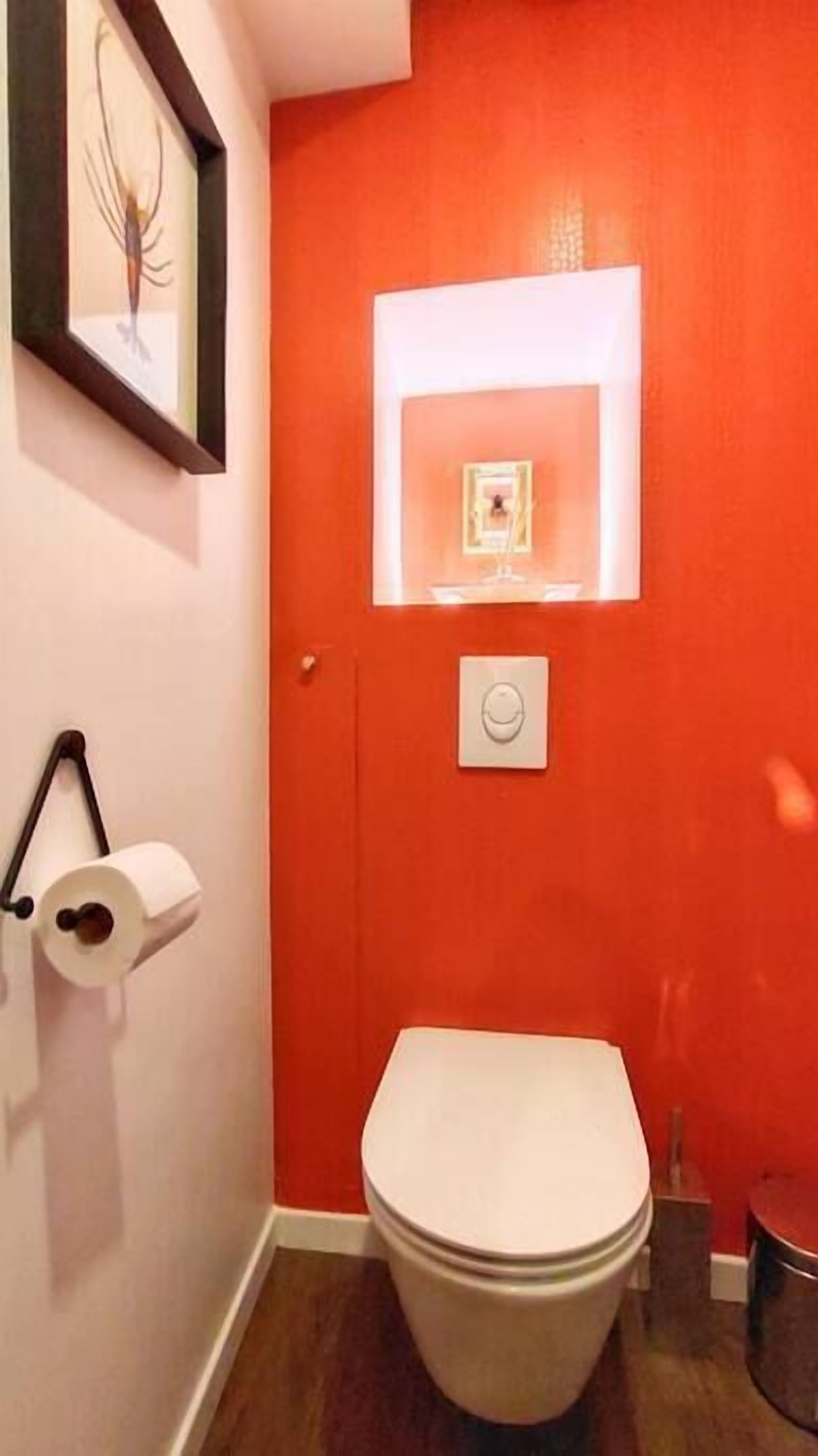 Une salle de bain avec des toilettes blanches, un rouleau de papier toilette, une œuvre d’art encadrée accrochée au mur et un mur d’accent de couleur rouge.