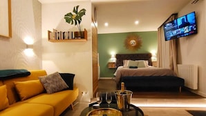 A hotel room with a yellow sofa, a bed with a dark headboard, a television on the wall, and a small table with a remote control and glasses.