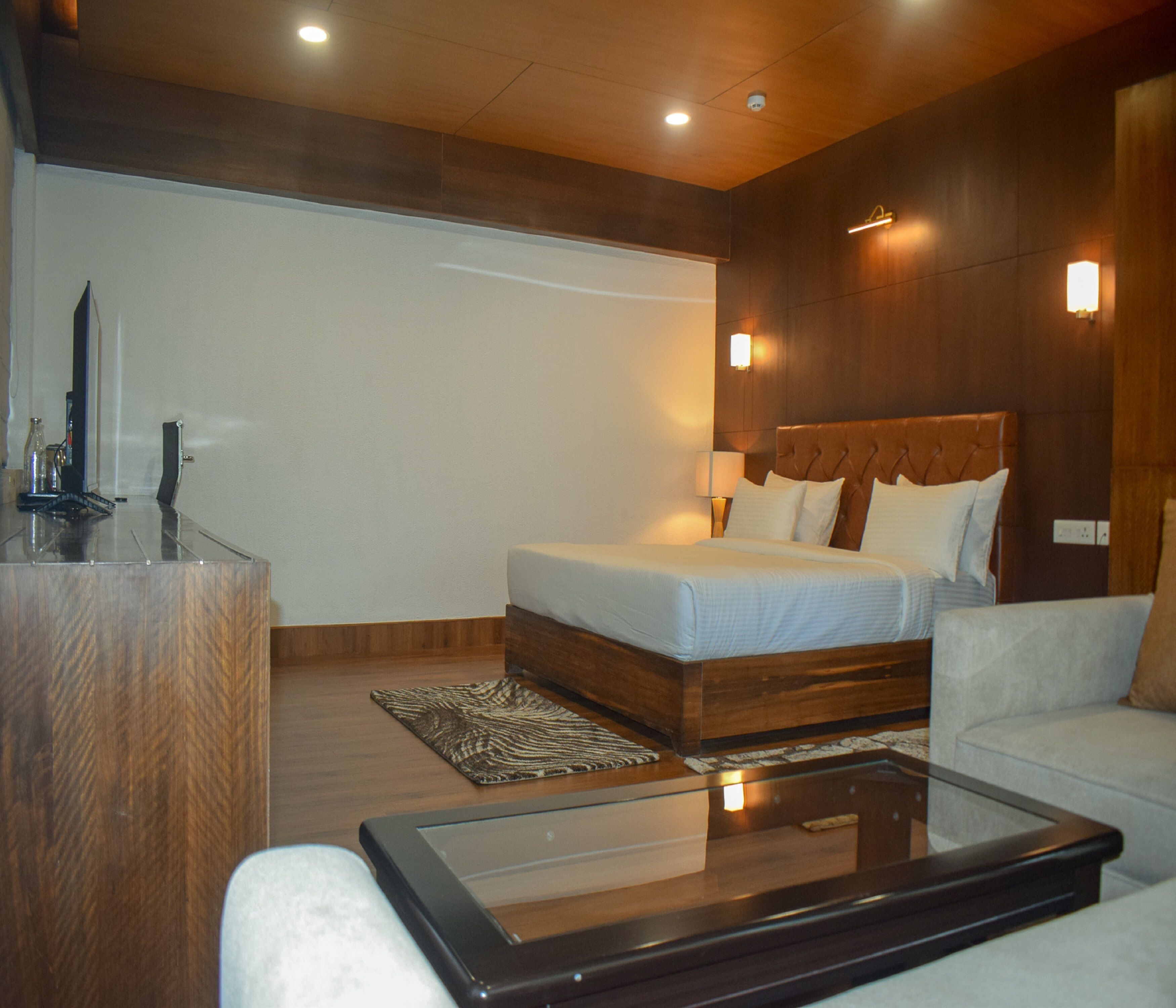 A modern hotel room with a large bed, a glass coffee table, a wooden desk, and a sofa.