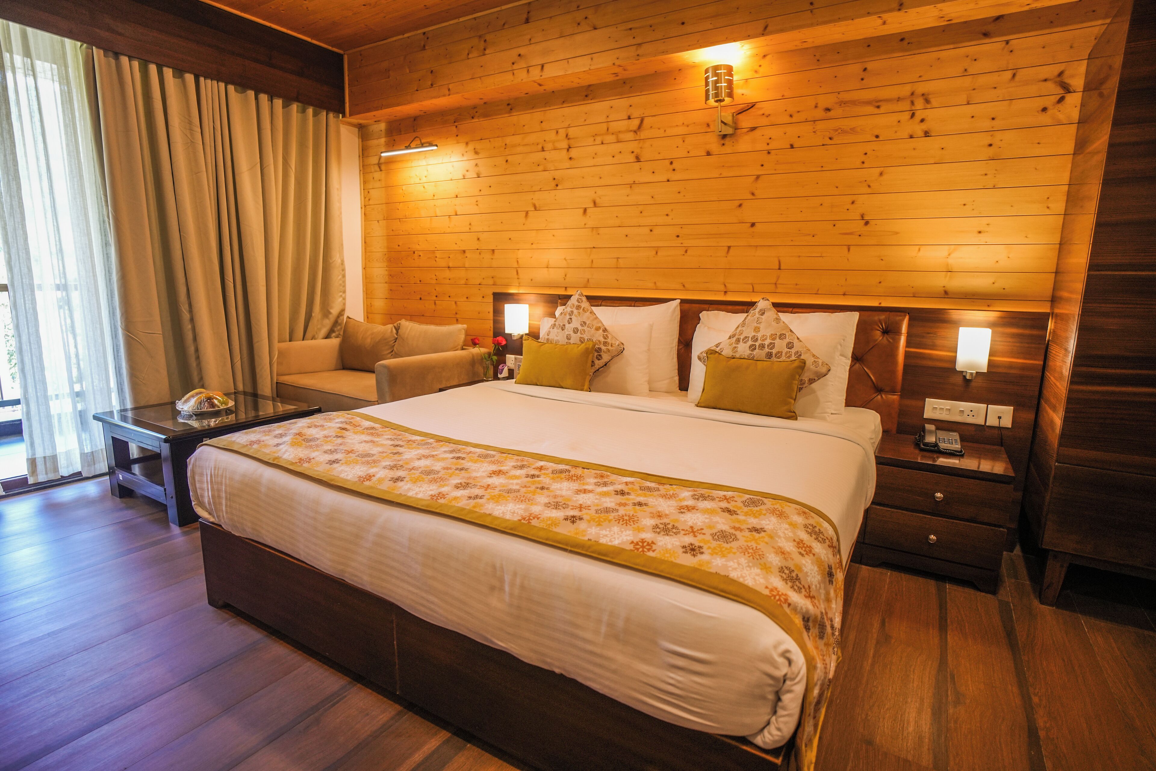 Quarto de hotel com teto de madeira, uma cama grande com colcha florida, duas mesas de cabeceira e um criado-mudo com luminária, além de uma janela com cortinas.
