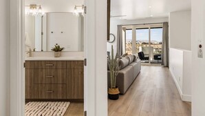 A modern bathroom with a wooden vanity, a mirror, and a potted plant. The bathroom leads to a living area with a sofa, a coffee table, and a view of the outdoors. - Waters Edge by RedAwning (St. George)