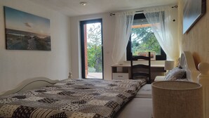 Slaapkamer met een bed, een bureau en een stoel. Er is een raam met gordijnen, een schilderij aan de muur en uitzicht op bomen.