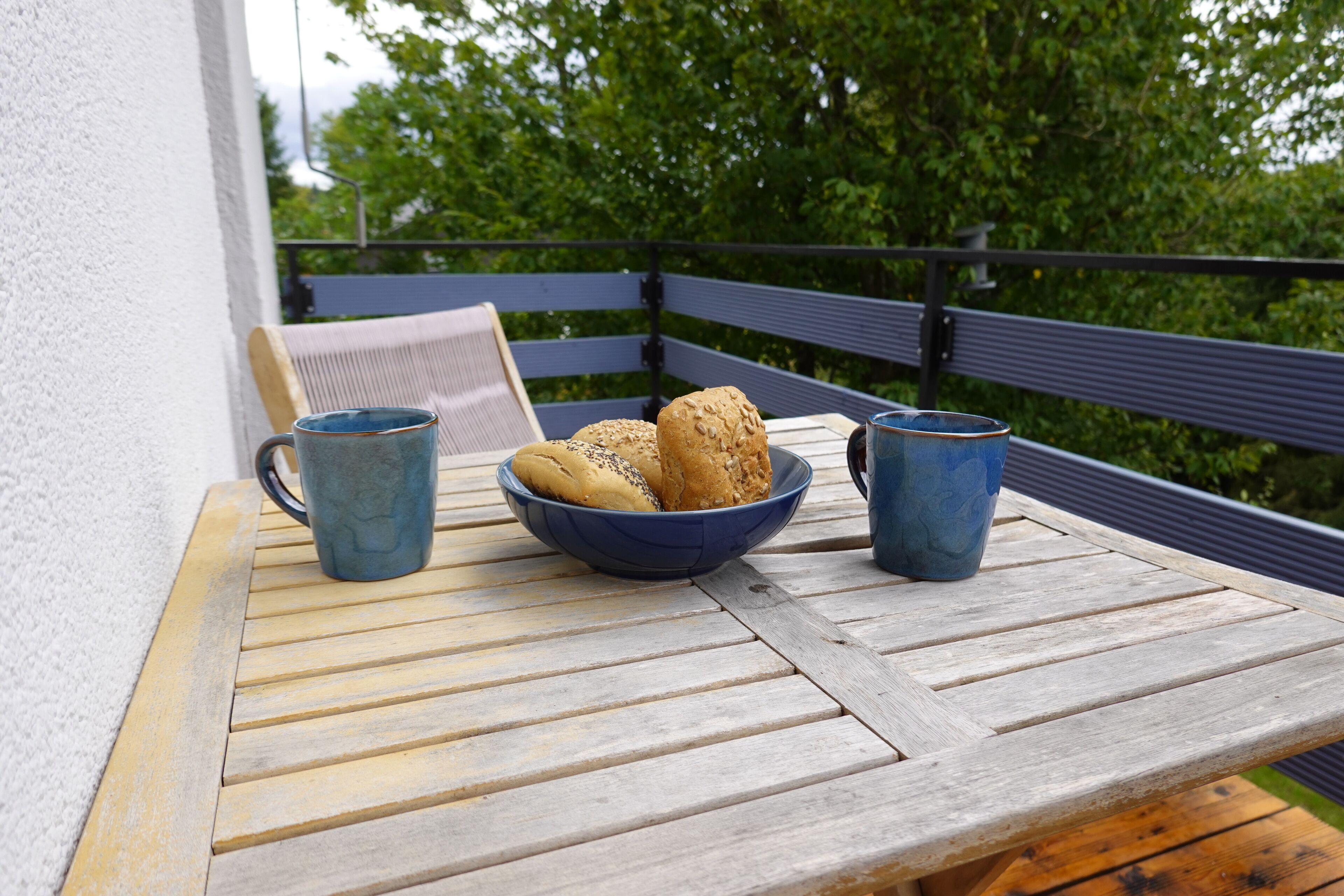 A wooden table with a bowl of bread and two blue mugs, set on a balcony with a view of trees.