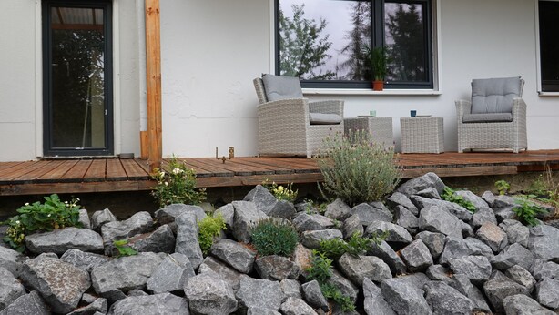 Een modern huis met een houten terras, twee rieten stoelen en een groot raam met uitzicht op bomen.