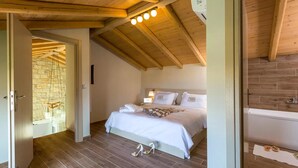 Quarto com teto de madeira, cama com roupa de cama branca, parede de pedra e chão de madeira.