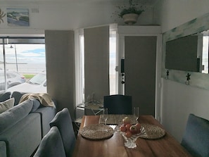 Un comedor con una mesa de madera, individuales tejidos y una superficie de vidrio. Se observa el exterior a través de una puerta, y hay una planta en el techo.