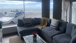 Un salón con un sofá gris, una mesa de centro con una planta en maceta, y vistas al mar a través de amplios ventanales.