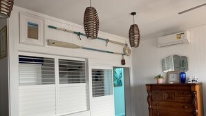 Una habitación con paredes blancas empañadas, un aparador de madera con cajones, un reloj y un espejo. Hay dos lámparas colgantes de mimbre, una ventana con persianas y una puerta que da a otra habitación.