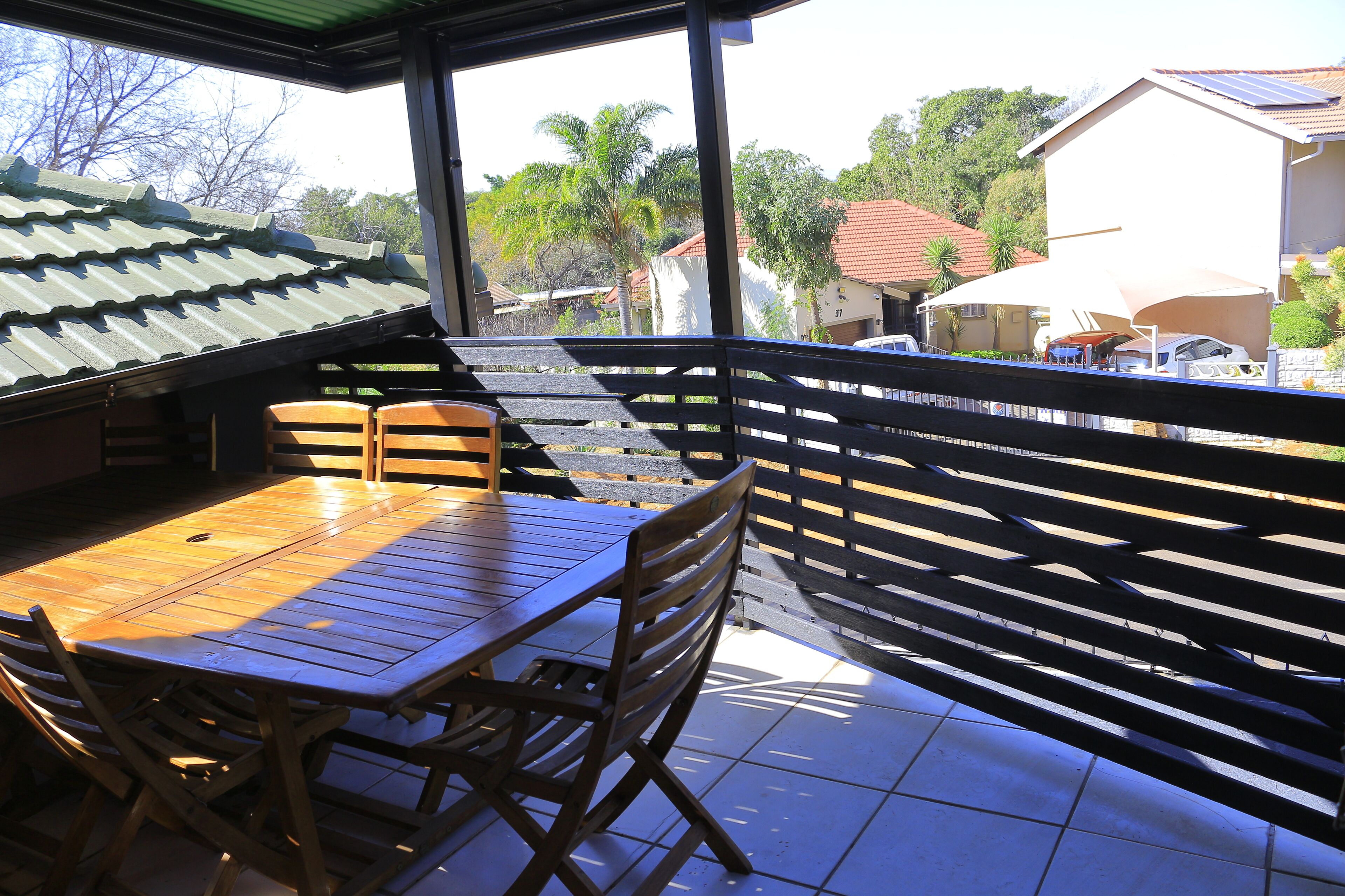 A wooden table and chairs on a balcony with a metal railing, overlooking a residential area with houses and trees.