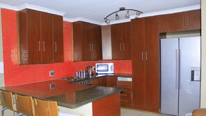 A kitchen with wooden cabinets, a stainless steel refrigerator, a microwave, and a bar area with stools. - Novel Enterprises Bed and Breakfast (Rustenburg)