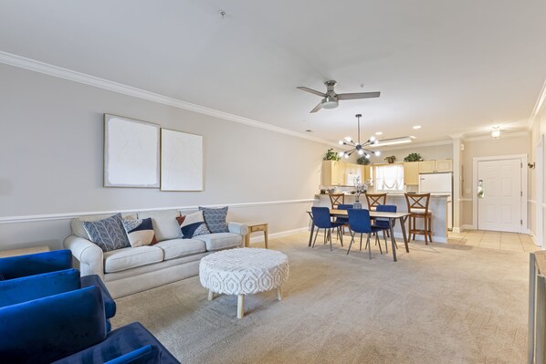 A living room with a sofa, ottoman, and blue chairs, a dining area with a table and chairs, and a kitchen in the background.