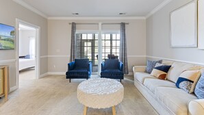 A living room with a beige carpet, a round ottoman, a white sofa, two blue armchairs, and a large window with curtains. - Stylish Golf  Beach Getaway (Myrtle Beach)