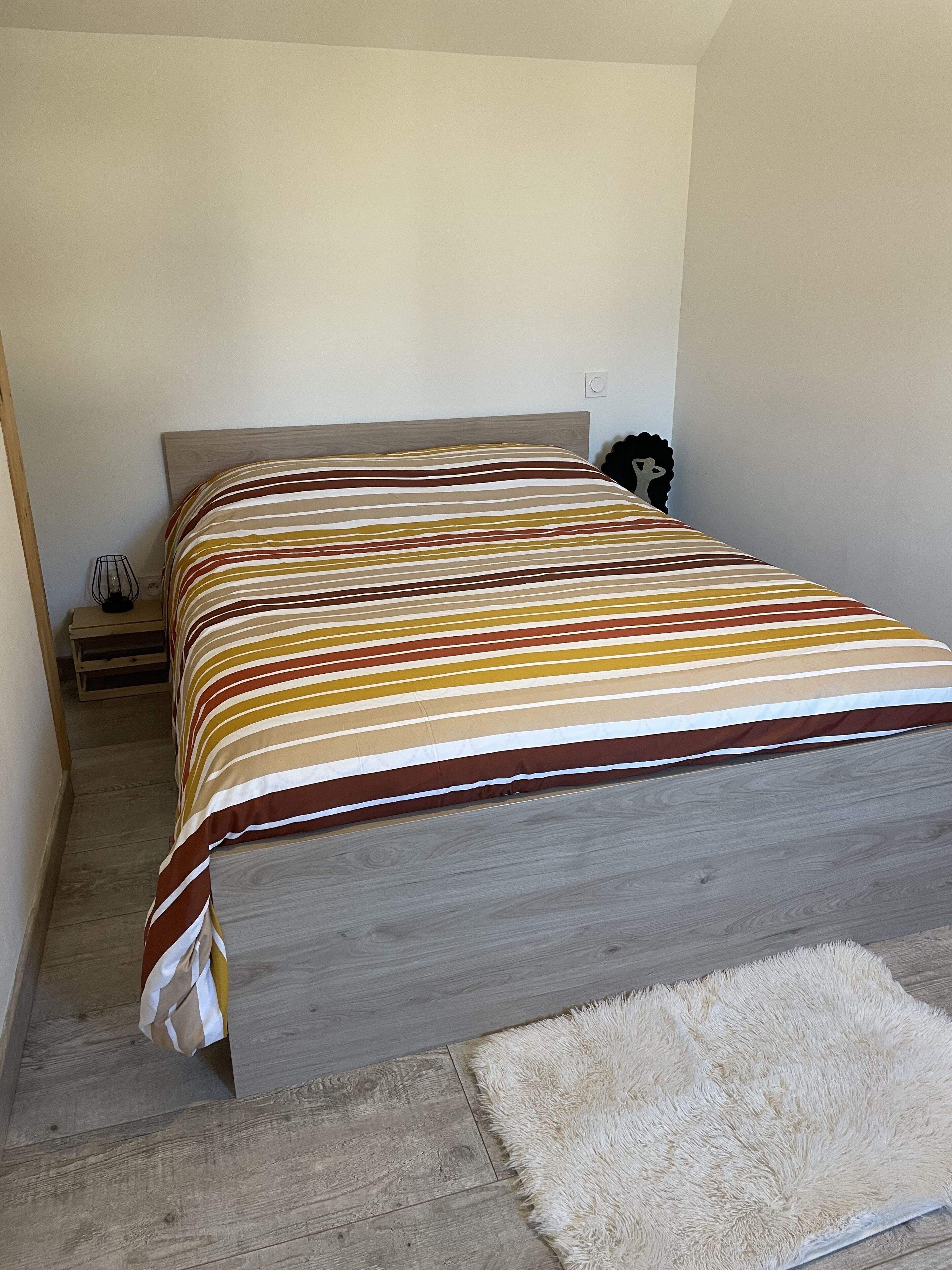 A bed with a striped bedspread, a wooden headboard, a bedside table with a lamp, and a fluffy rug on the floor.