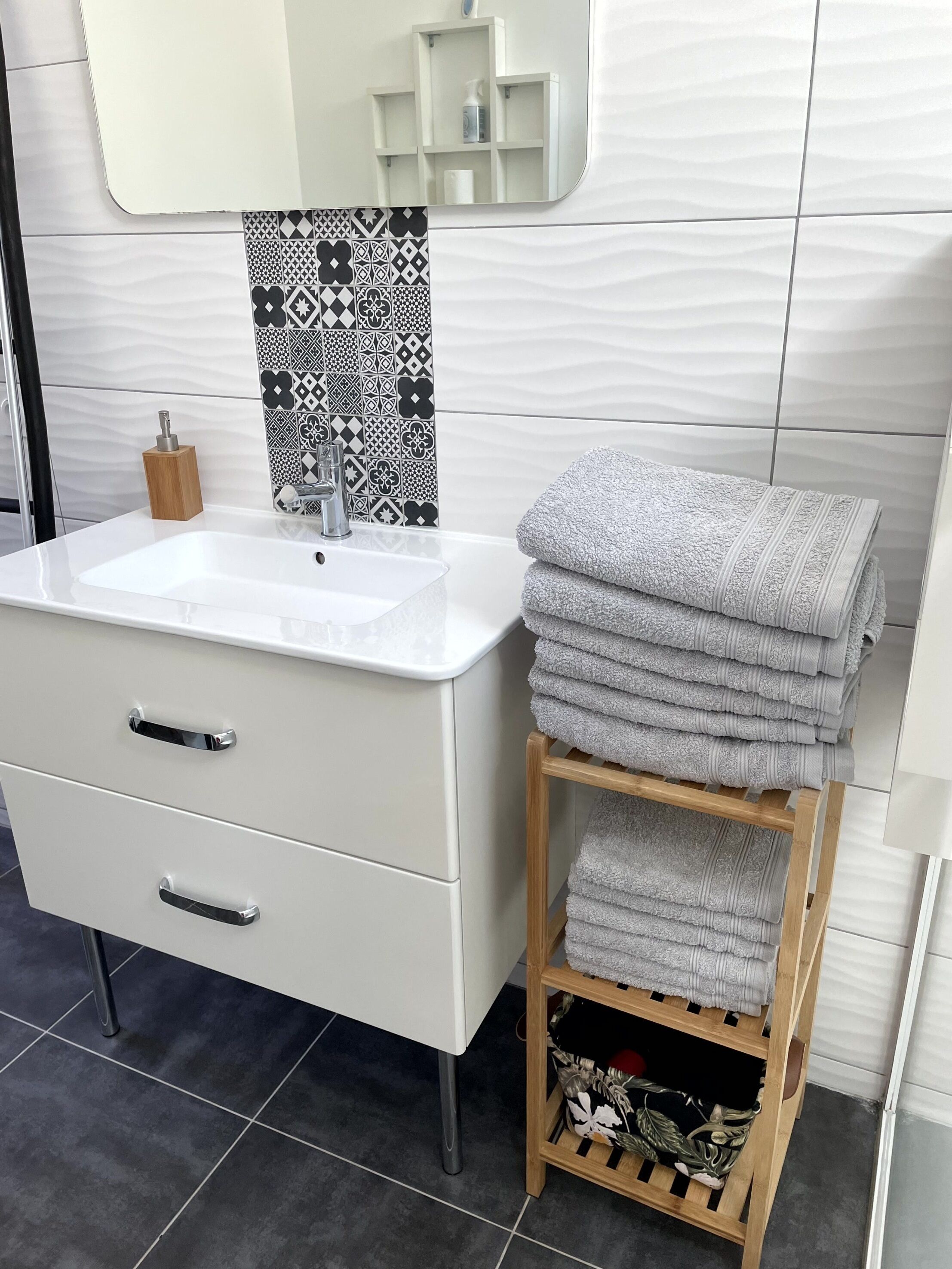 A bathroom with a white sink, a mirror, a patterned tile backsplash, and a wooden towel rack with folded towels.