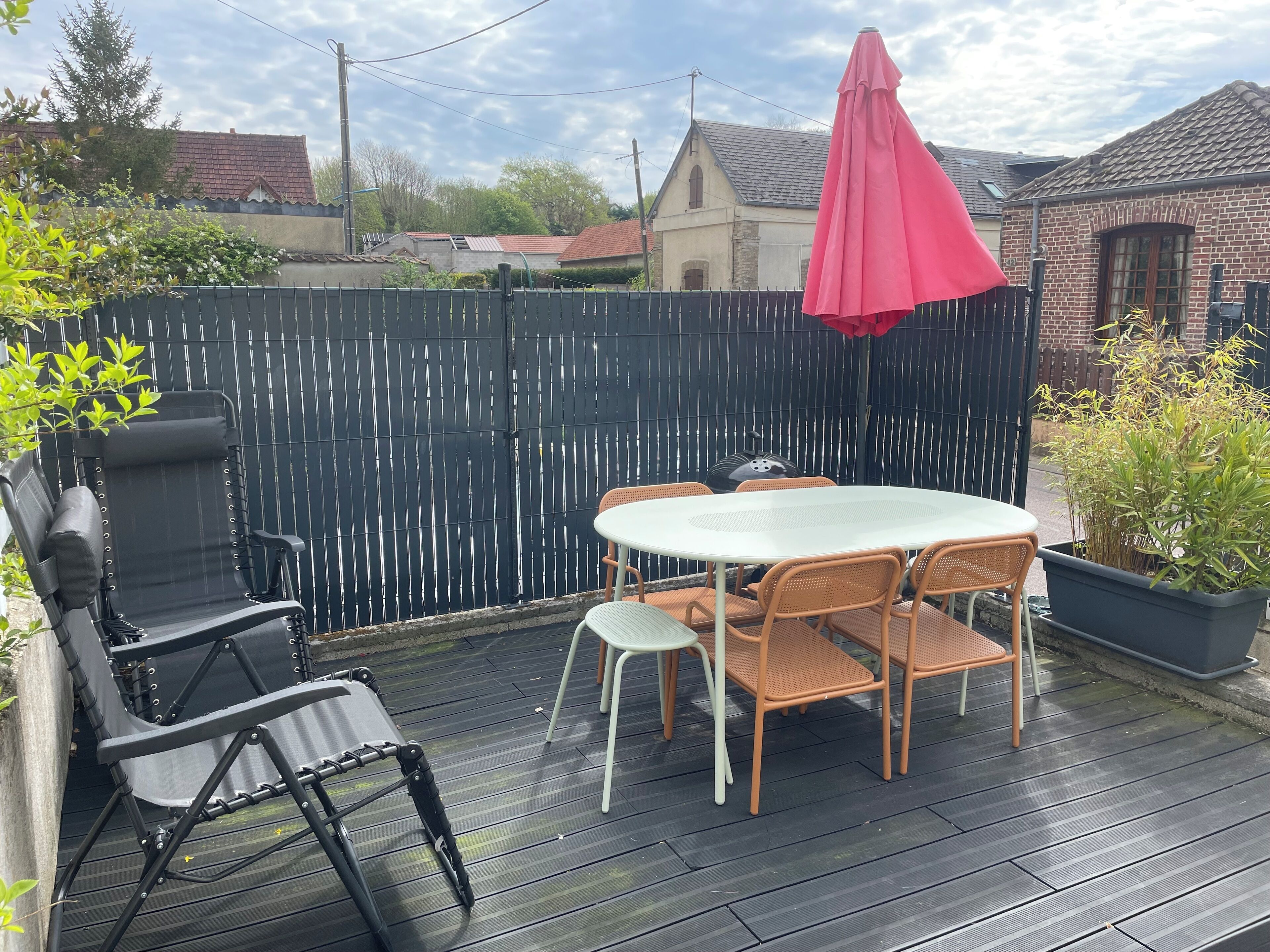 Une terrasse avec une table ronde et des chaises, une chaise pliante noire, un parasol rouge et une terrasse en bois.