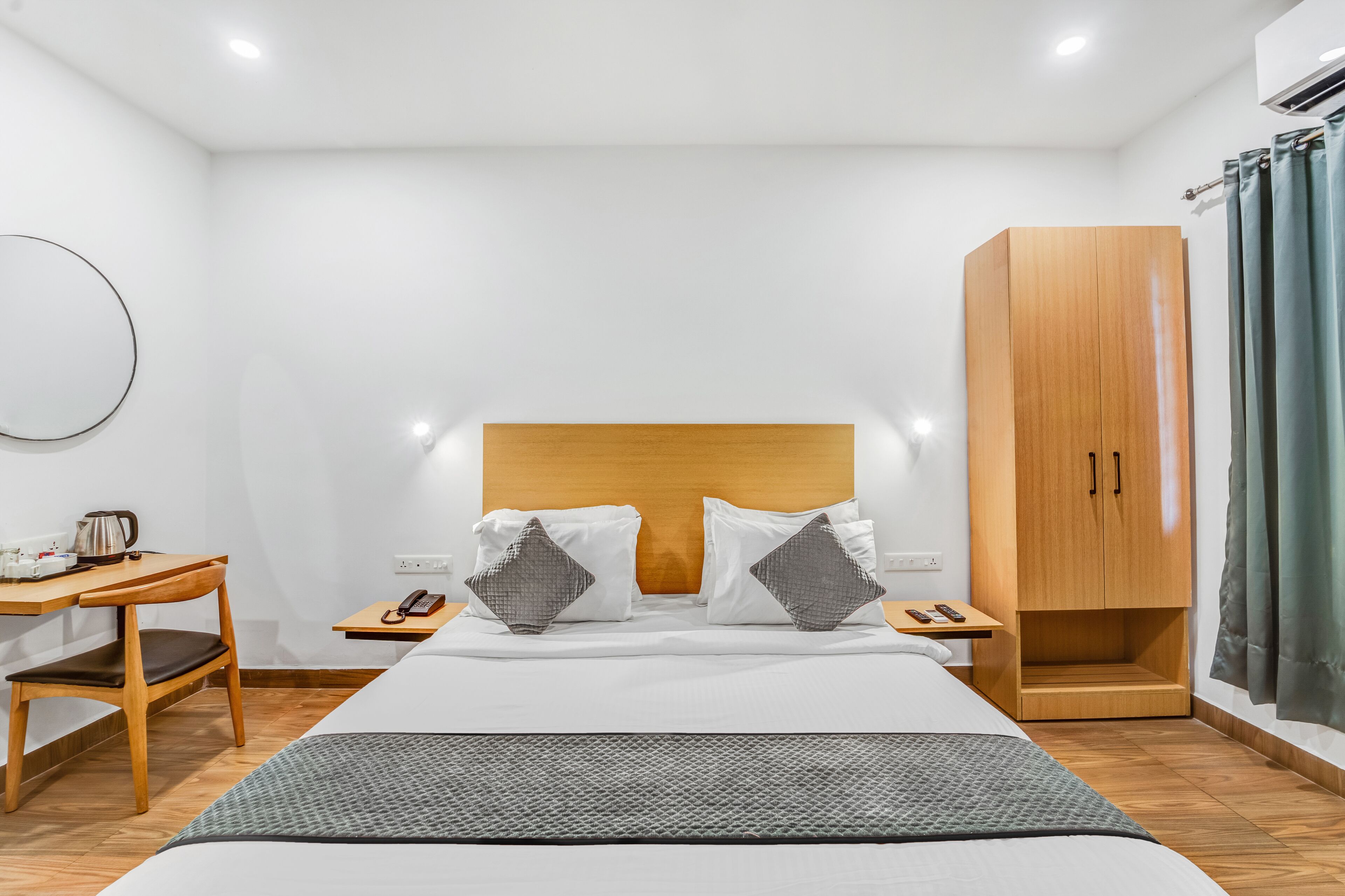 A hotel room with a large bed, wooden headboard, a desk with a chair, a wardrobe, and a small table with a phone and a kettle.