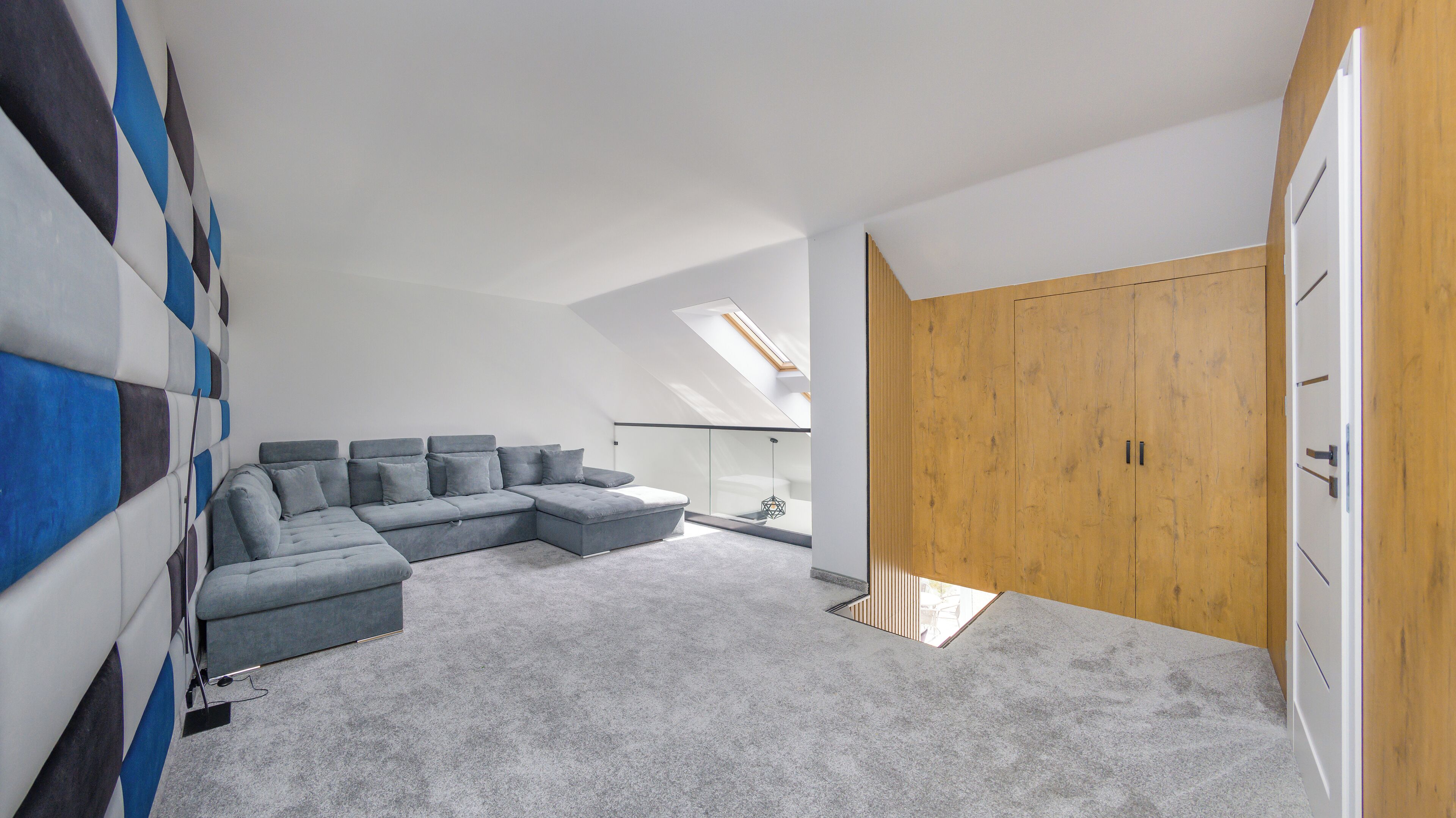 A modern loft bedroom with a large L-shaped sectional sofa, a skylight, and a wooden wardrobe.