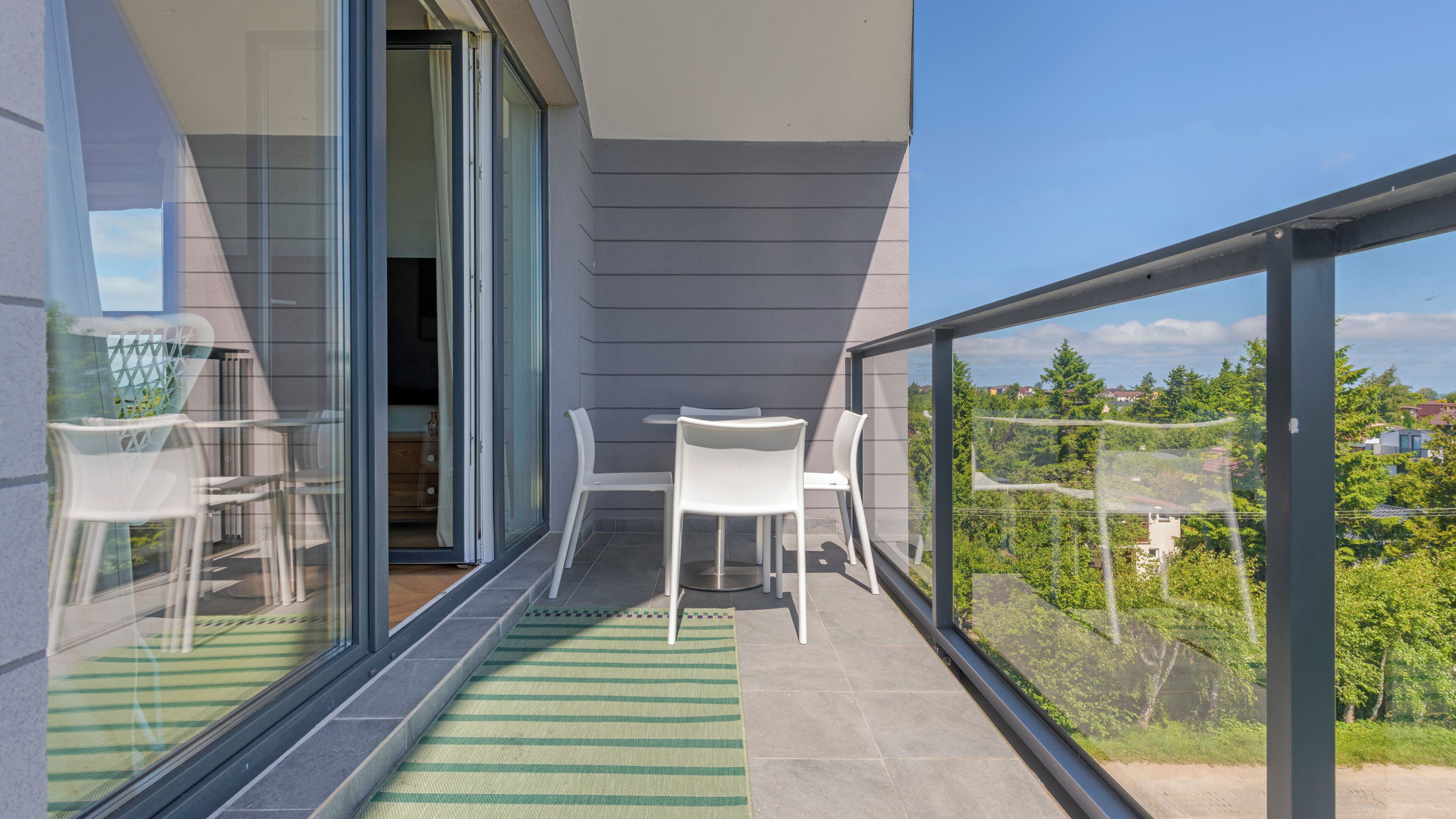 Un balcon avec une porte vitrée, des chaises blanches, une petite table et un tapis à rayures vertes.
