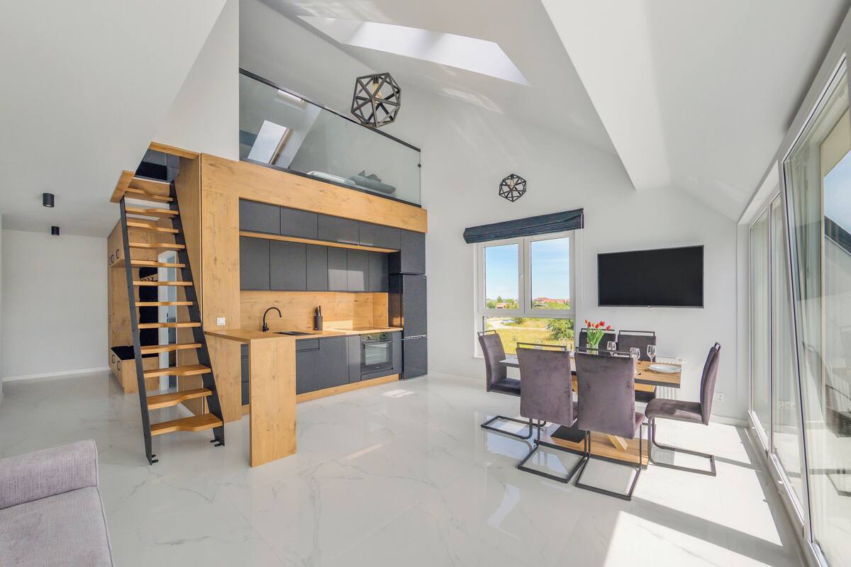 A modern kitchen with a wooden staircase, a dining area with a table and chairs, and a large window offering a view.
