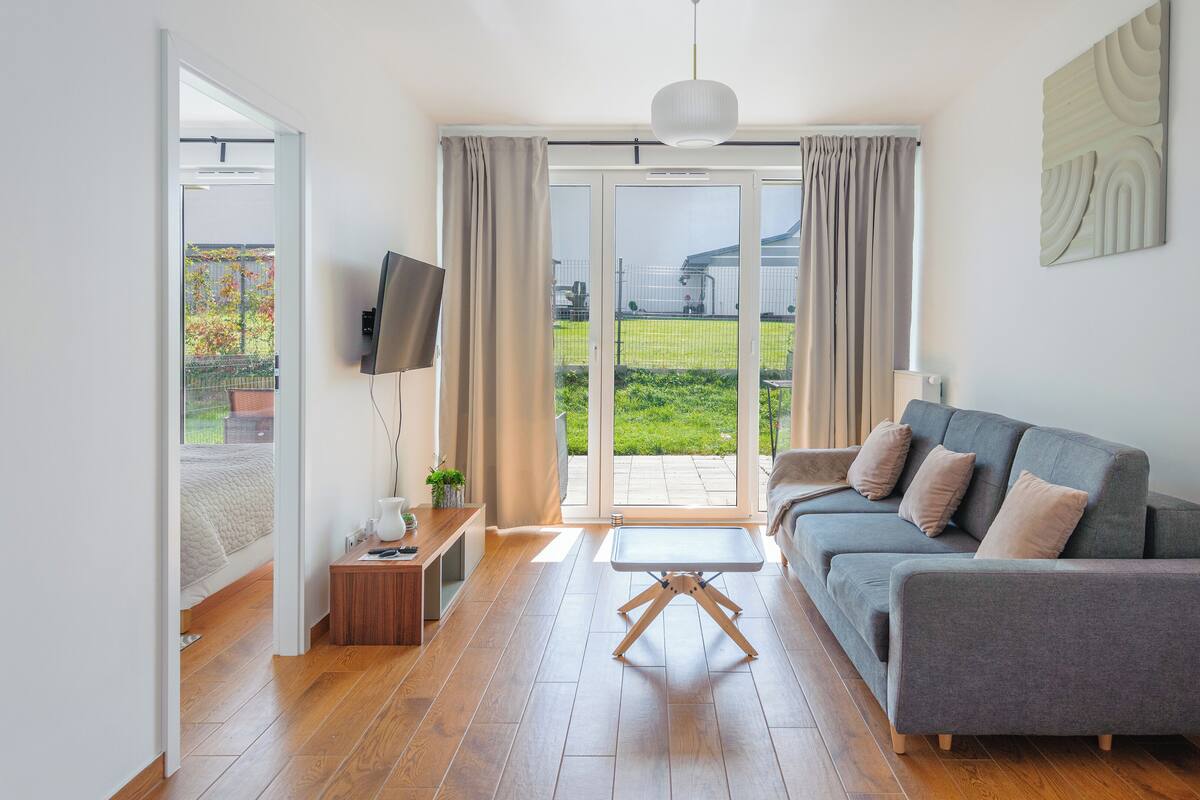 A modern living room with a grey sofa, wooden flooring, and a view of a green garden through a sliding glass door.