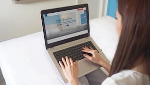 Uma pessoa usando um laptop na cama, que possui um colchão branco, com uma parede azul ao fundo.