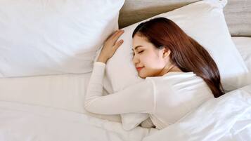 A person lying in bed with white bedding and pillows, in a room with a wooden headboard.
